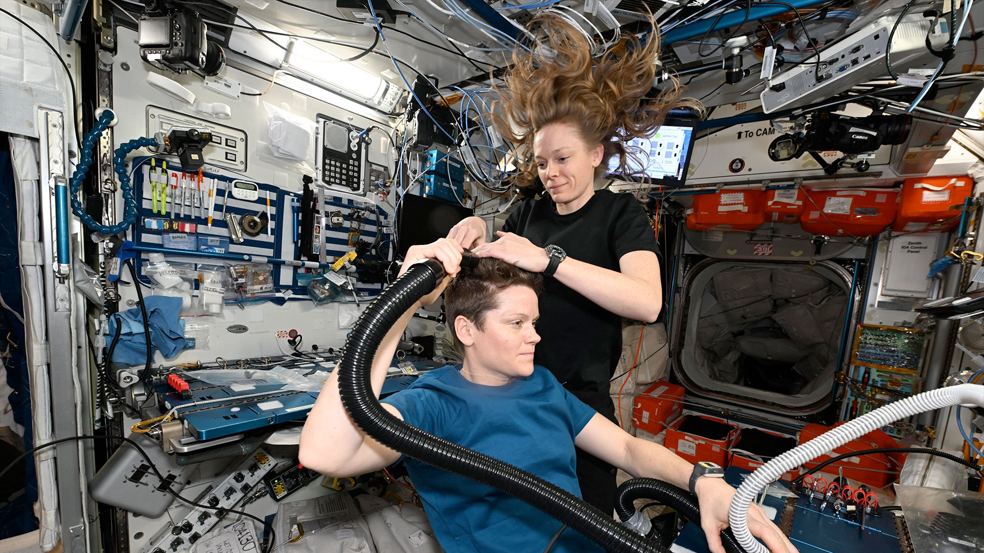 a woman in a dark blue shirt gives a haircut to another woman in a lighter blue shirt aboard a space station