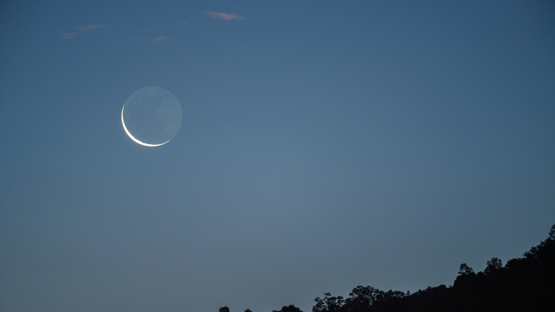 A waning crescent moon is pictured rising in the morning sky above a dark horizon.