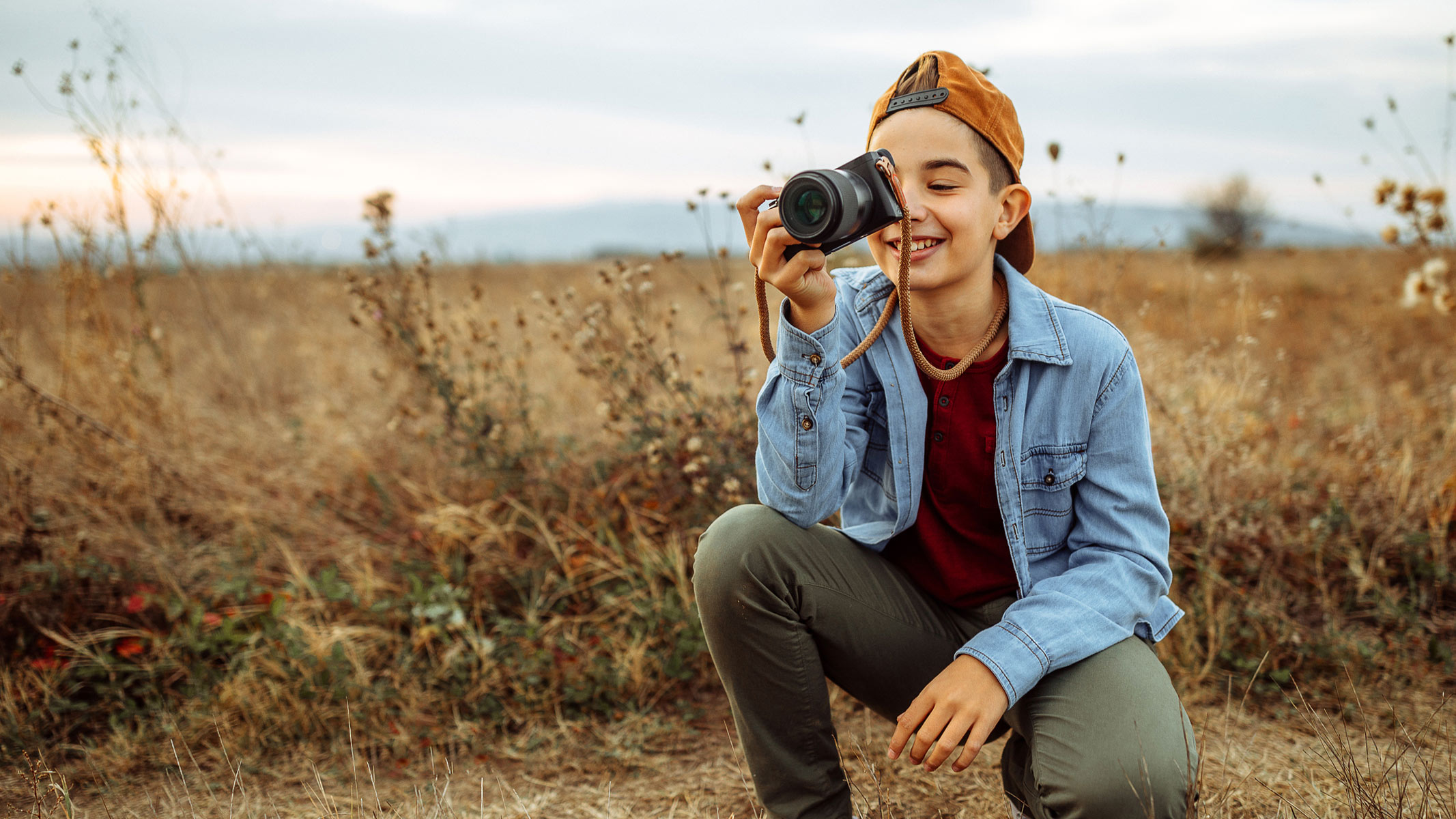 A teenager with a cap holding a camera near their face kneeling in a brown field.