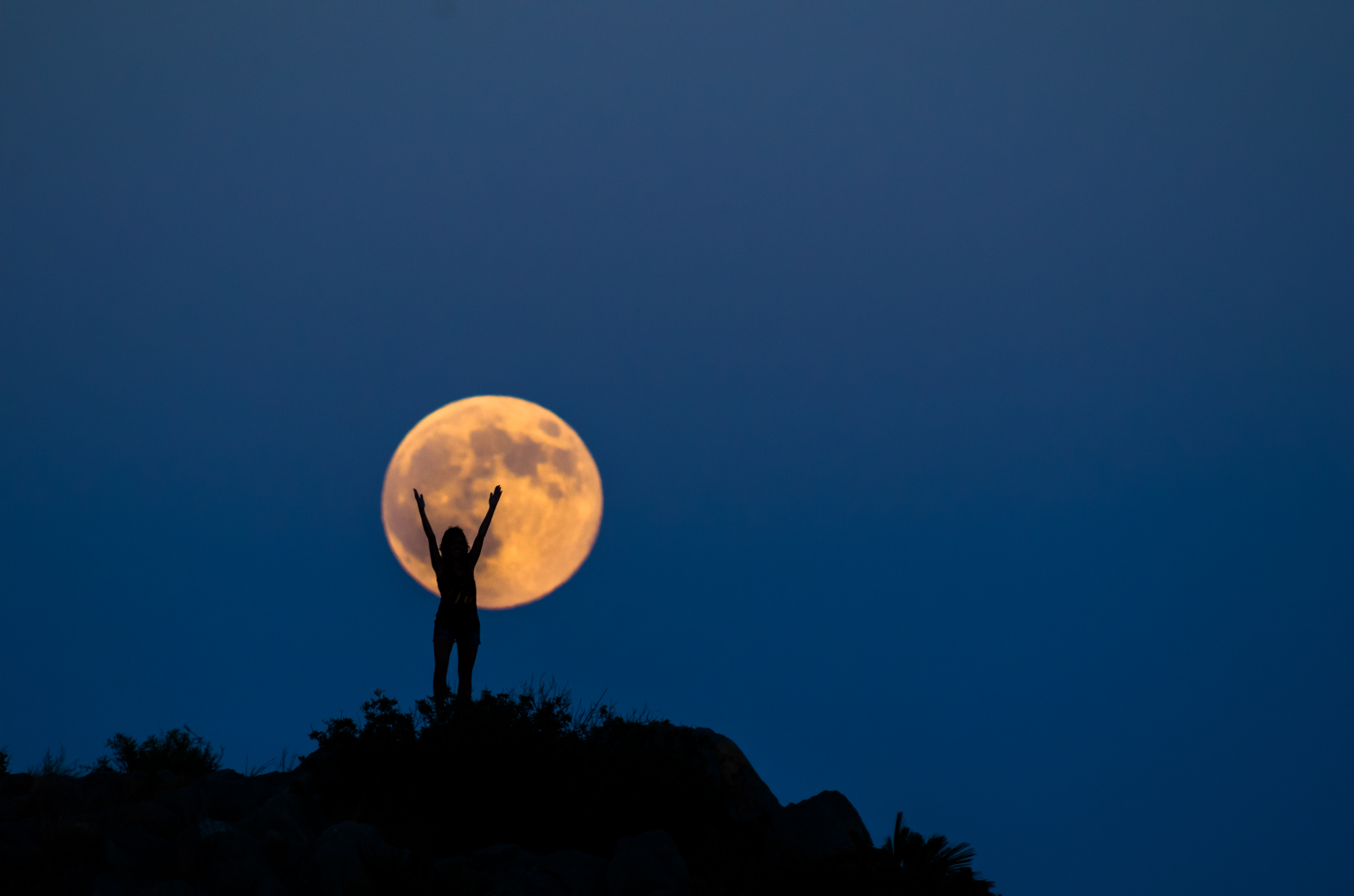 A woman with raised arms is silhouetted against a yellow full moon, which sits close to the horizon against a dark blue sky.