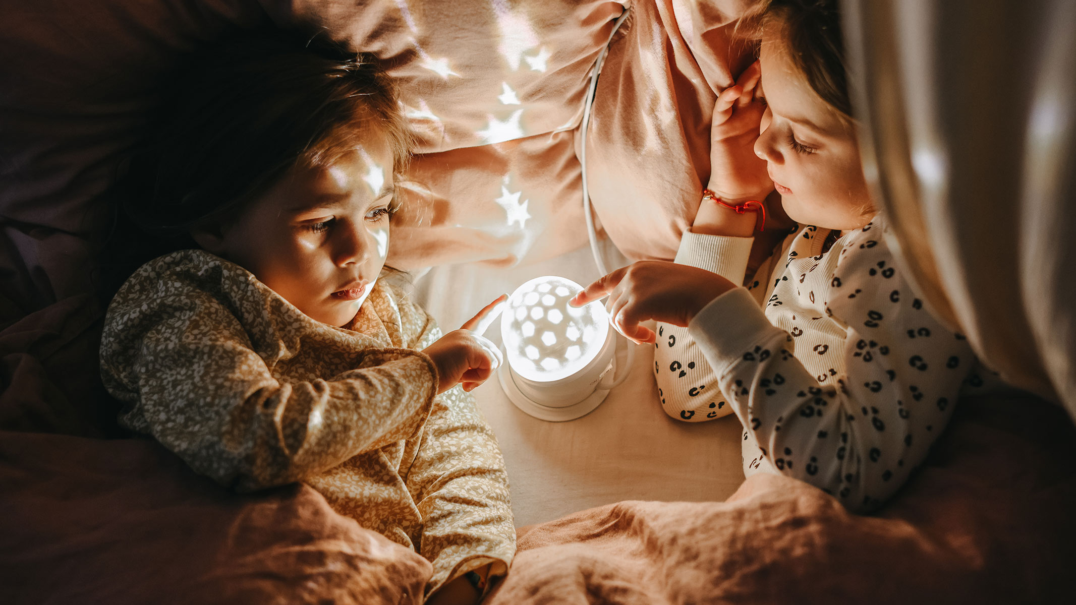 Two children in bed placing their finger on a star projector
