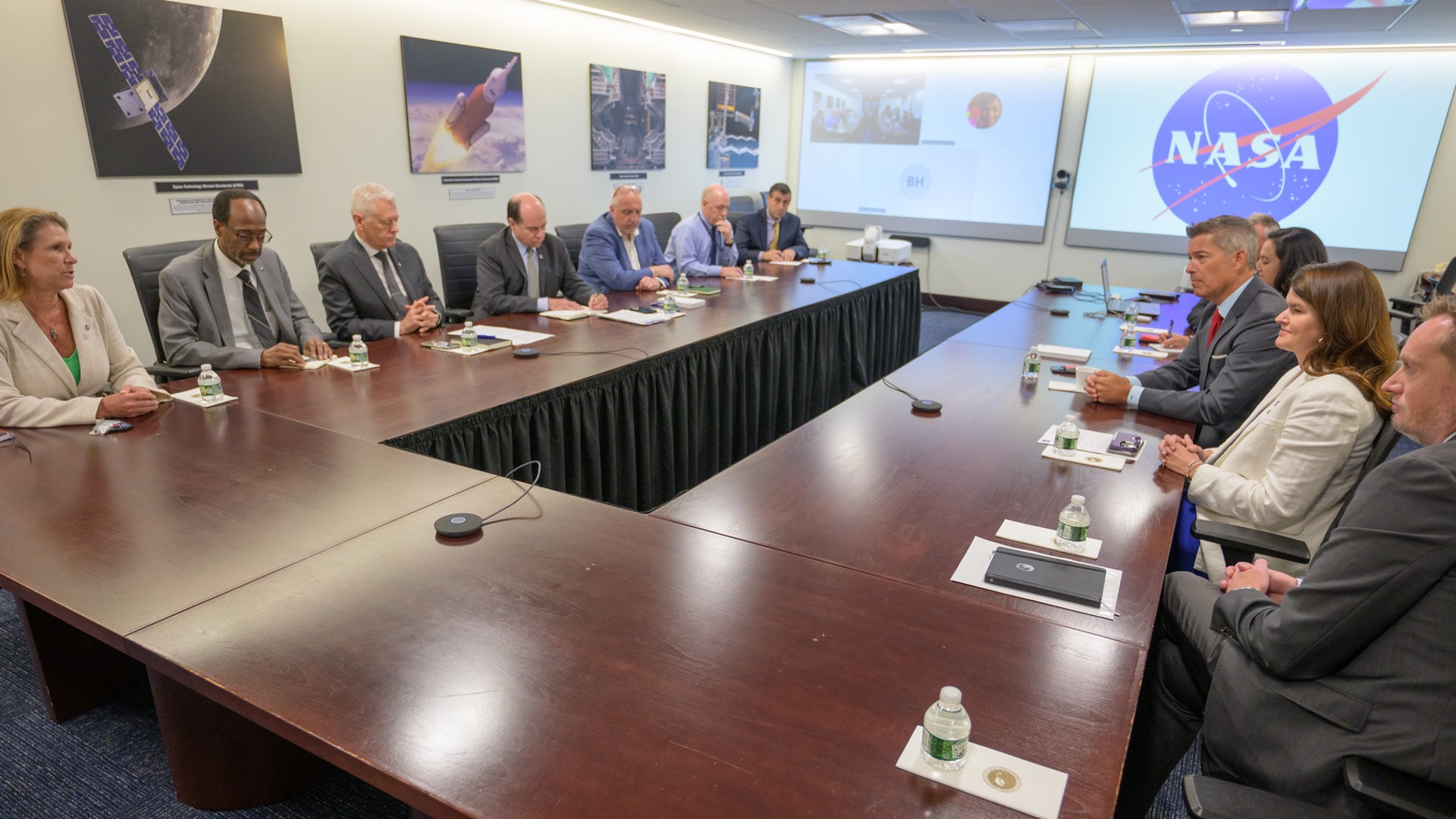 a group of people in business attire sit at a C-shaped table in front of a NASA logo