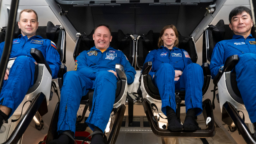 Four humans in blue jumpsuits sit in a spacecraft.