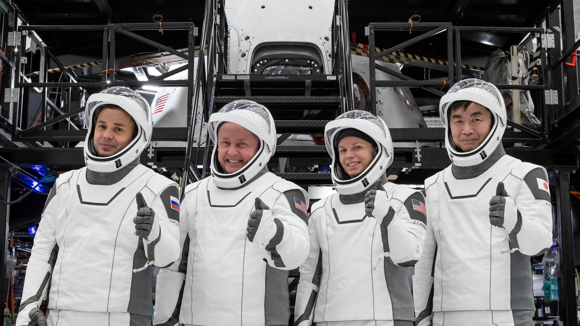four people in white spacesuits smile and give a thumbs-up in front of a cone-shaped white spacecraft in a hangar surrounded by scaffolding