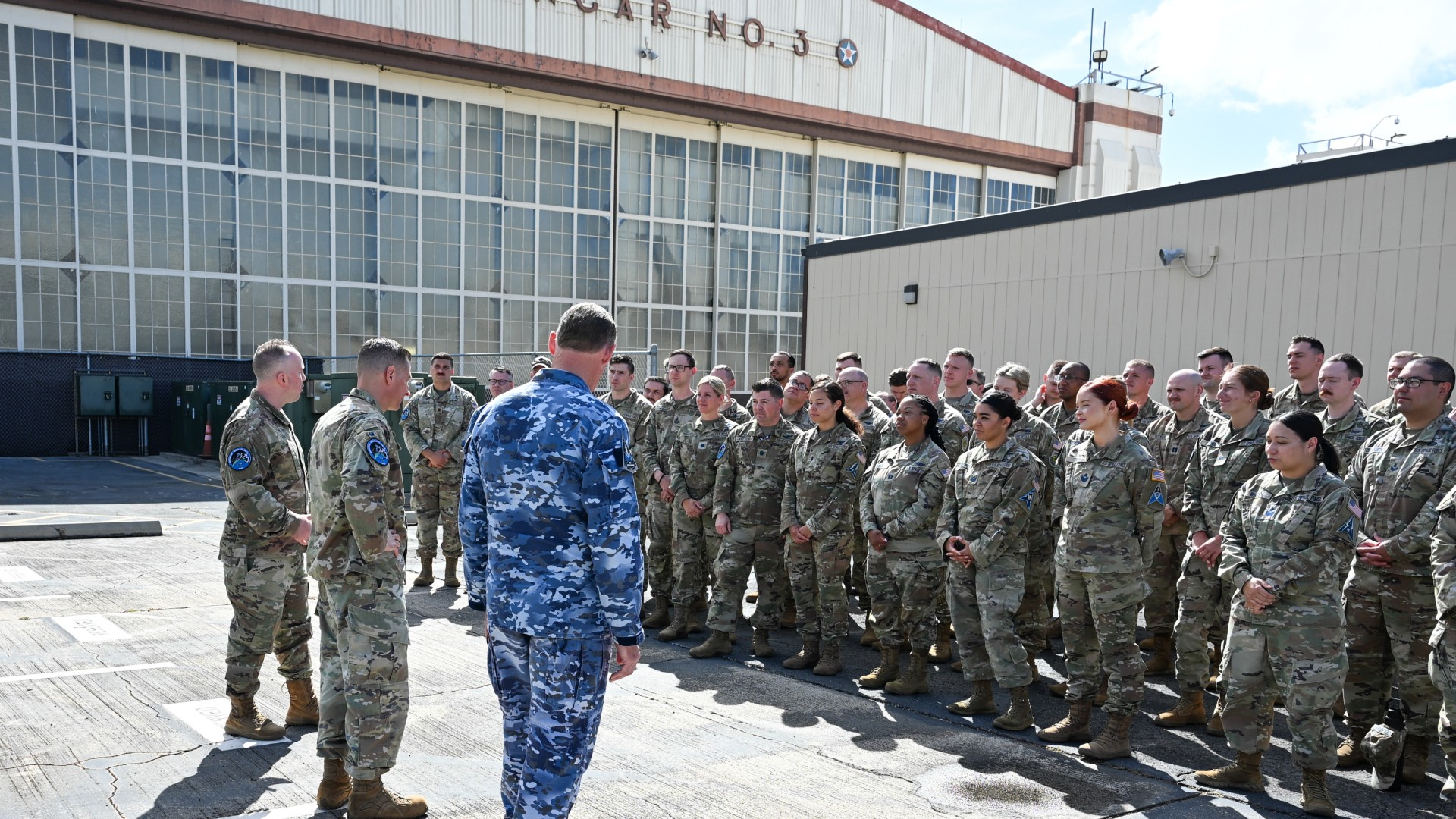 three men in camouflage uniforms address a crowd of people also in camouflage uniforms outside a hangar on a concrete tarmac