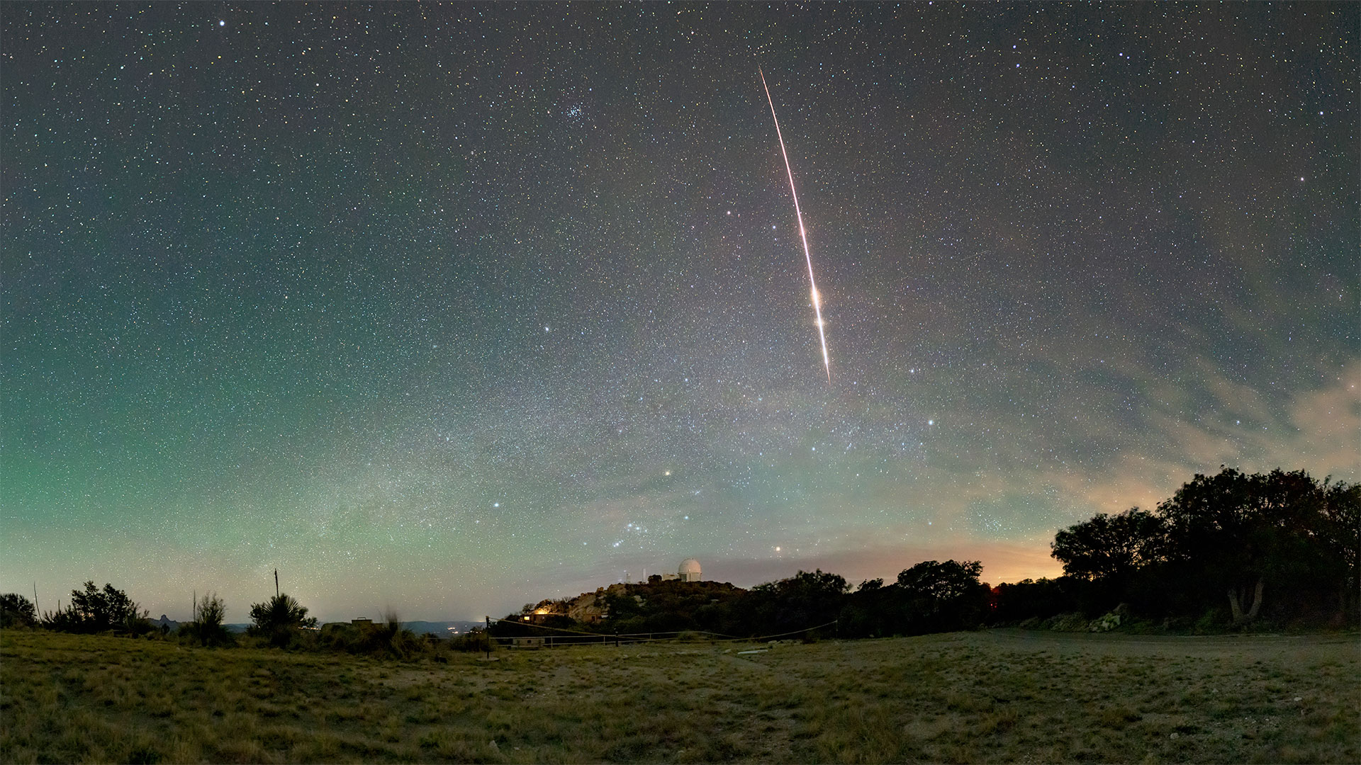 a red-tinted streak from a meteor plunges through a greenish-blue sky above the dome of an astronomical observatory