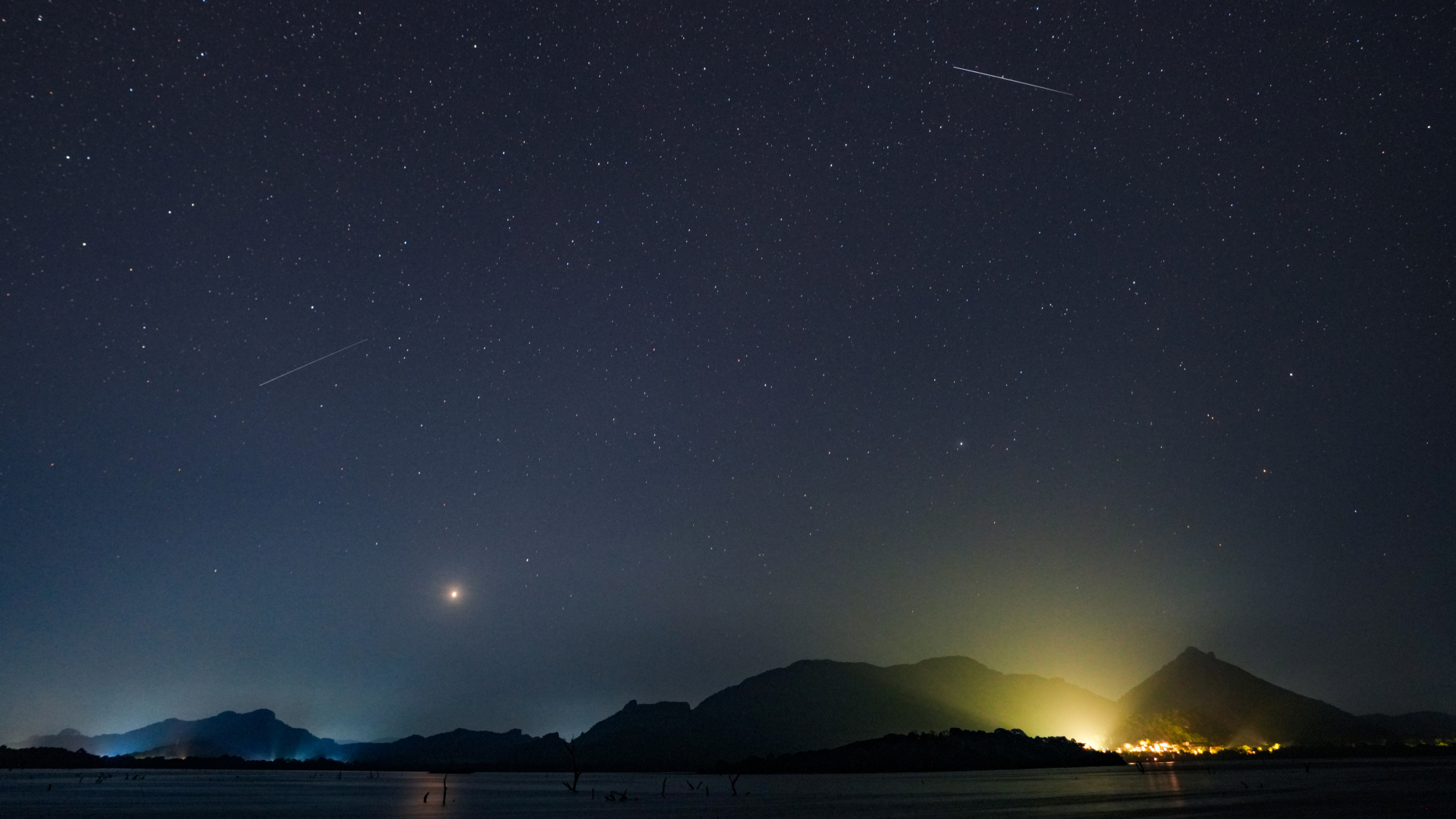 Two Eta Aquarid meteors can be seen streaming across the star studded sky above Sri Lanka, Light from settlements bordering a body of water can be seen spilling into the sky.