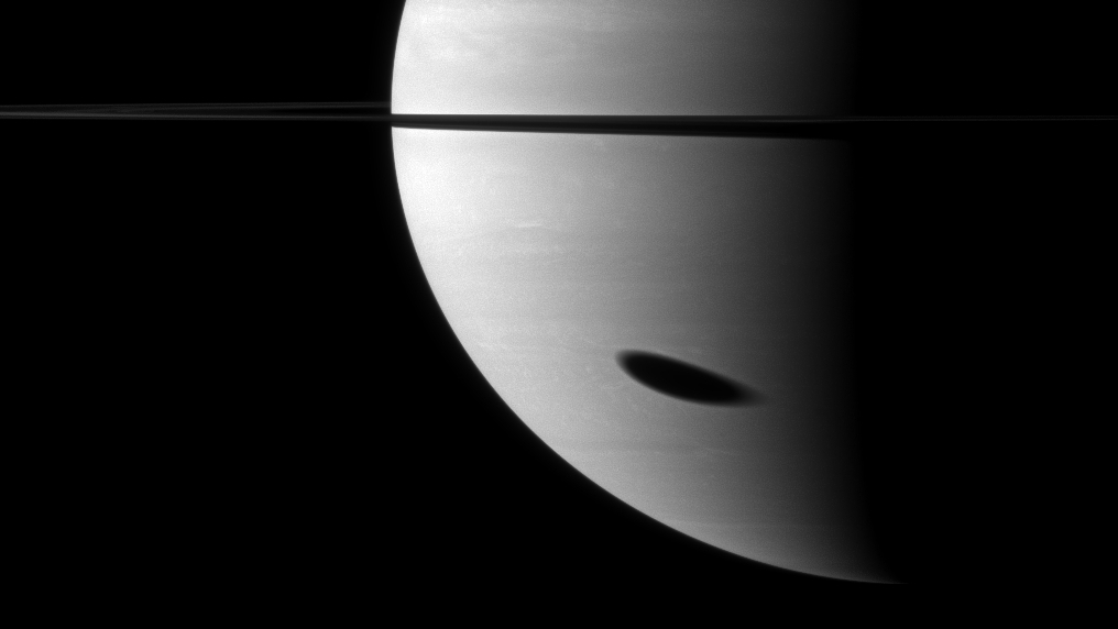 Saturn is pictured with its left half illuminated by the sun while its rings are edge on against a black background. The elongated shadow of the large moon Titan is visible on the lower half of the planet