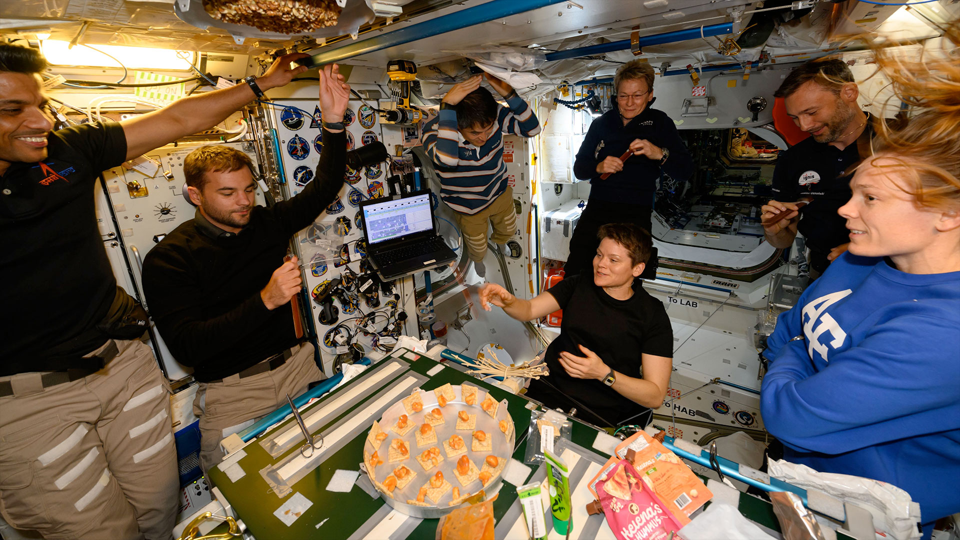 seven people in polo shirts and slacks gather around a table to enjoy a meal together aboard a space station